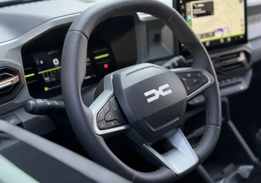 Vue rapprochée du volant noir du Dacia Duster 2025 avec commandes intégrées et écran tactile GPS en arrière-plan.