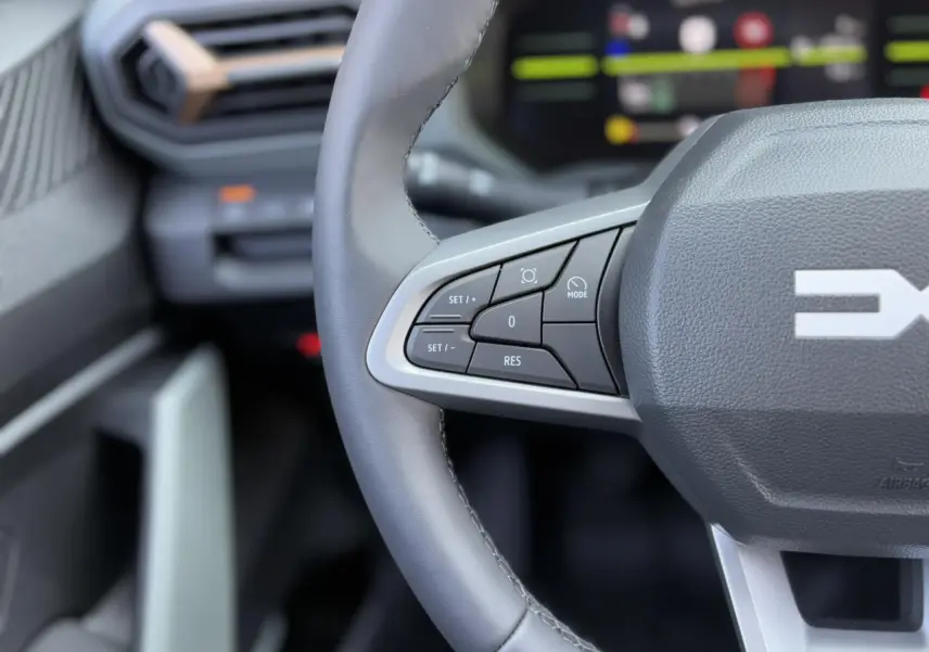 Gros plan sur le volant gris du Dacia Duster 2025 avec commandes intégrées et tableau de bord digital flou en arrière-plan.