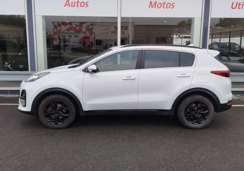 Profil côté gauche du KIA Sportage blanc 2022 avec jantes noires et vitres teintées, stationné devant un showroom.