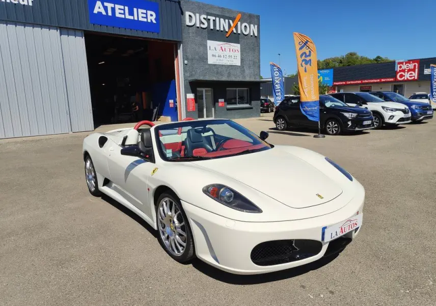 Ferrari F430 Spider blanc vue 3/4 avant droit, capote ouverte, intérieur cuir rouge, devant un garage.