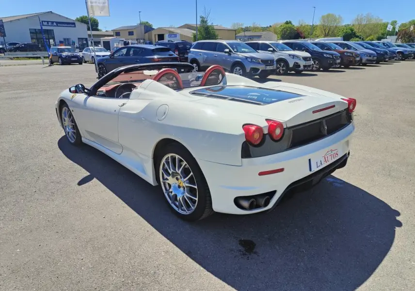 Ferrari F430 Spider blanc vue 3/4 arrière droit, capote ouverte et intérieur rouge visible, stationnée en extérieur.