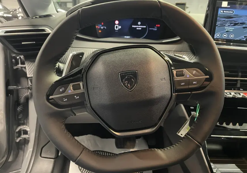 Vue rapprochée du volant cuir noir de la Peugeot 208 1.2i Hybrid 2025 avec commandes multimédia intégrées et tableau de bord numérique.