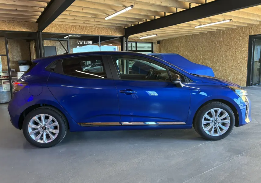 Profil droit d'une Renault Clio 2024 bleu iron, avec jantes alliage et détails chromés sous les portières, en intérieur d'atelier.