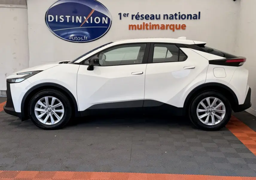 Profil côté gauche du Toyota C-HR blanc 2024 avec lignes anguleuses et jantes alliage visibles en intérieur showroom.