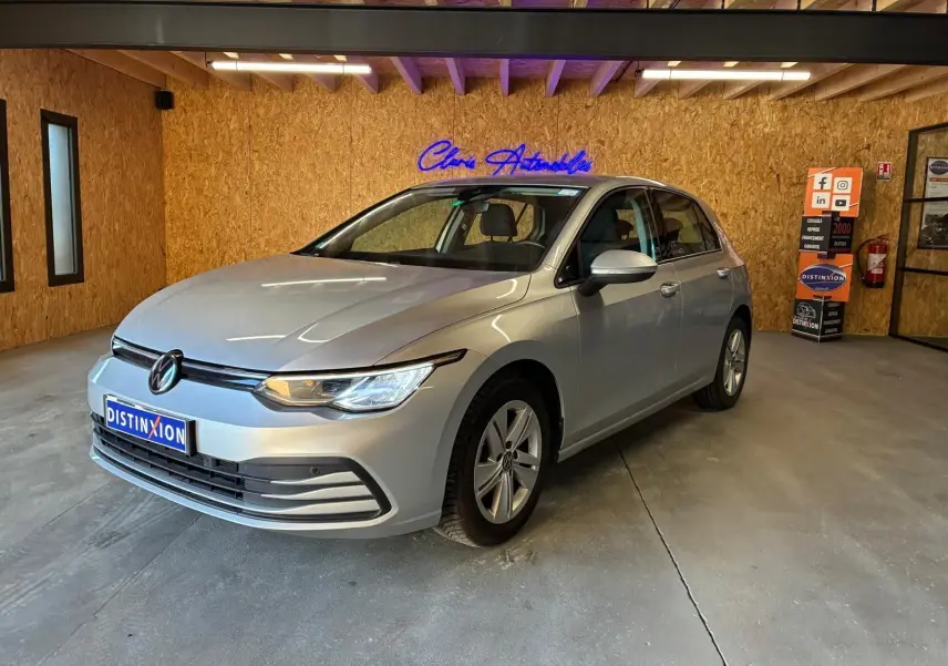 Volkswagen Golf gris clair vue 3/4 avant droit dans un showroom avec éclairage intérieur et panneau publicitaire.