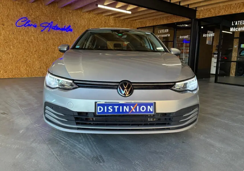 Vue avant d'une Volkswagen Golf 1.0 eTSI gris clair 2023 avec plaques Distinxion dans un showroom.
