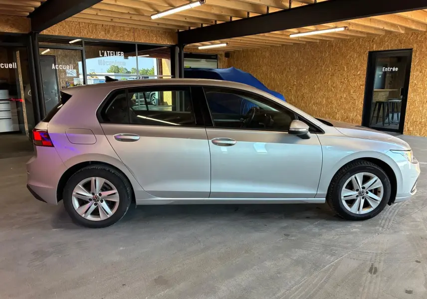 Vue de profil côté gauche d'une Volkswagen Golf 1.0 eTSI 110 DSG7 Life Plus gris clair, stationnée en intérieur.