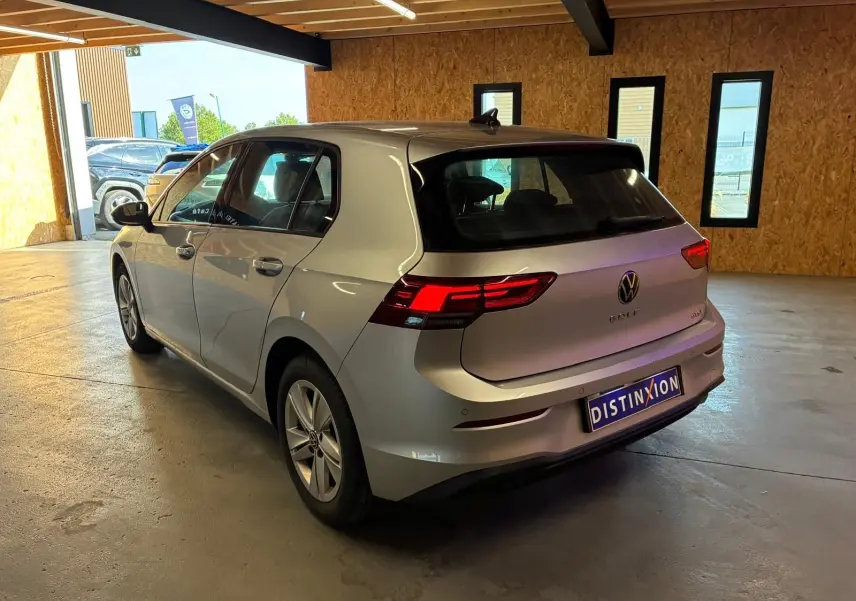 Volkswagen Golf gris clair 2023 vue 3/4 arrière droit avec feux arrière allumés en intérieur.