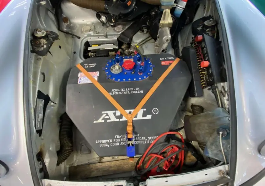 Vue plongeante sur le compartiment avant gris métallisé d'une Porsche 911 3.0 SC avec réservoir FIA noir et sangles orange visibles.
