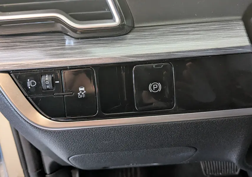 Gros plan sur les commandes noires du tableau de bord du Kia Sportage blanc MY25, boutons de réglage lumière et frein à main électronique.