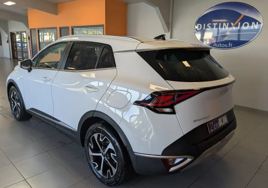 Vue 3/4 arrière droite du KIA SPORTAGE blanc 2025 avec jantes alliage bicolores et barres de toit en intérieur showroom.