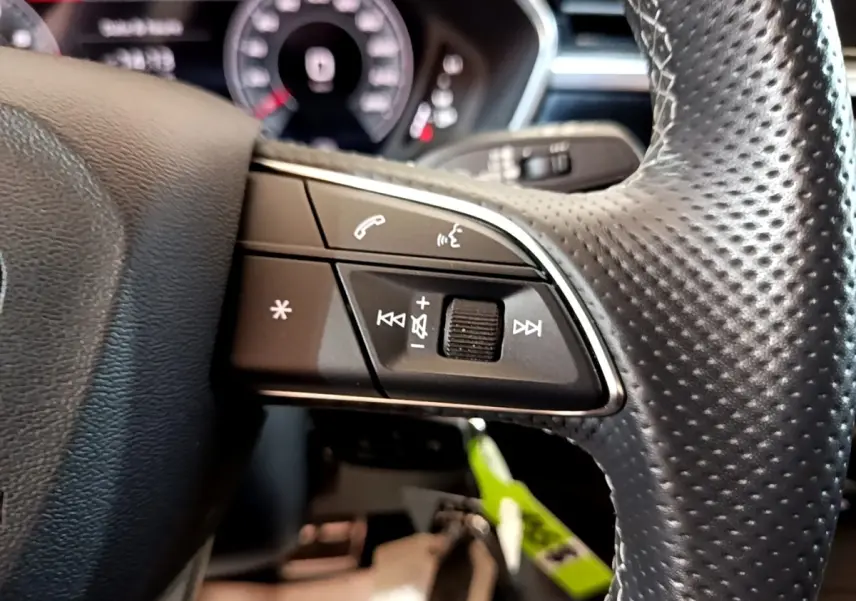 Gros plan sur les commandes gauche du volant cuir perforé de l'Audi Q3 gris foncé, avec tableau de bord numérique flou en arrière-plan.