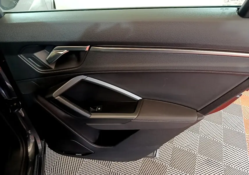 Détail de la porte intérieure côté gauche de l'Audi Q3 gris foncé avec poignée argentée et commande de vitre.