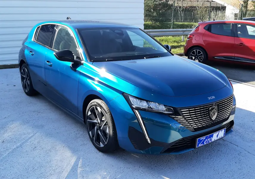 Vue frontale d'une Peugeot 308 Bleu Obsession 2026 avec calandre noire et logo lion distinctif, stationnée devant un mur blanc.