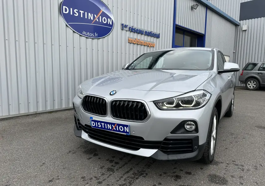 BMW X2 gris vue 3/4 avant avec calandre noire et phares LED allumés devant un bâtiment industriel