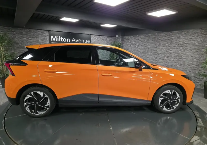 Profil côté gauche d'une MG4 électrique orange avec toit noir et jantes alliage bicolores dans un showroom.