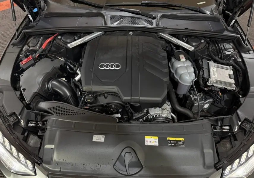 Vue plongeante sur le moteur noir d'une Audi A4 gris foncé, avec capot ouvert et logo Audi visible.