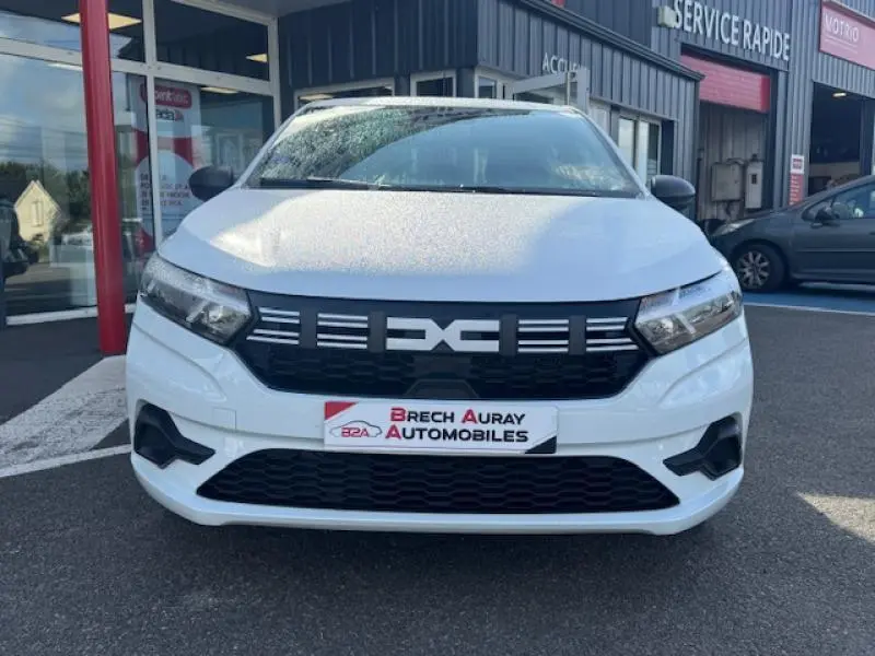 Vue avant d'une Dacia Sandero blanche 2024 avec calandre noire et phares LED, devant un garage automobile.