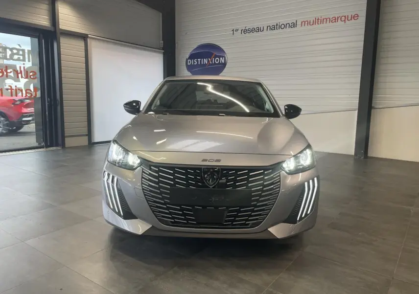 Vue frontale d'une Peugeot 208 1.2i Hybrid 2025 gris Artense avec phares LED allumés et calandre noire distinctive.