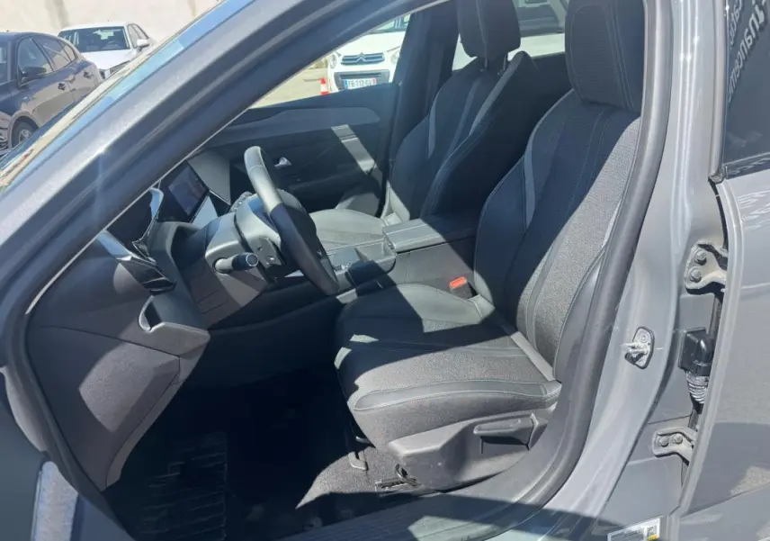 Intérieur avant de la Peugeot 308 Hybrid gris, vue côté conducteur avec sièges en tissu et accoudoir central visible.