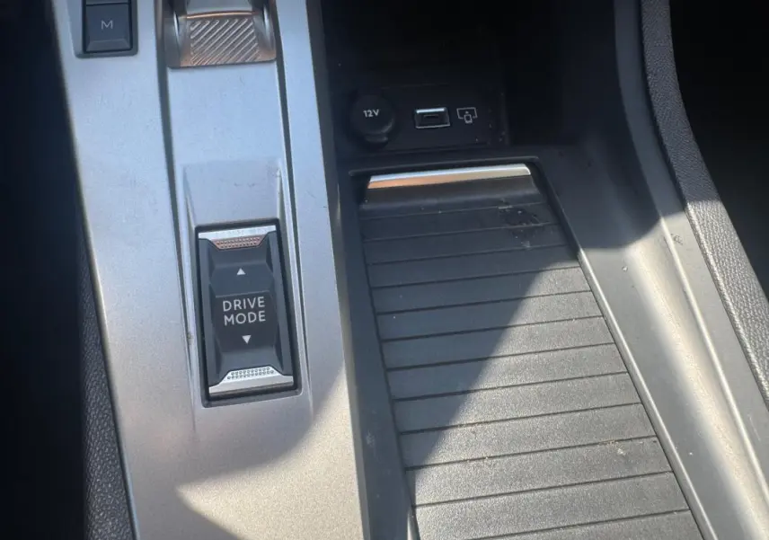 Gros plan sur la console centrale grise de la Peugeot 308 Hybrid 2025 avec bouton Drive Mode et prise USB visible.