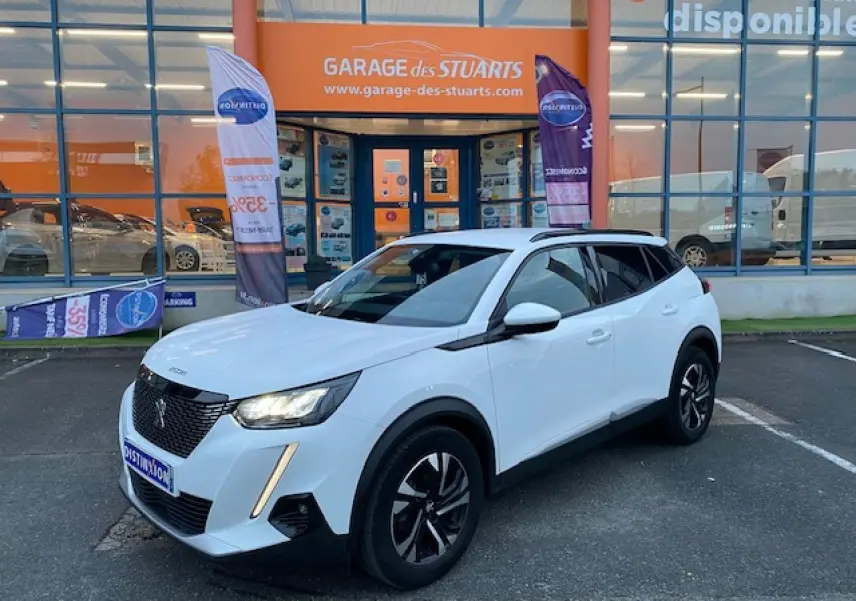 Peugeot 2008 blanc vue 3/4 avant droit, jantes alliage bi-ton et calandre noire devant un garage.