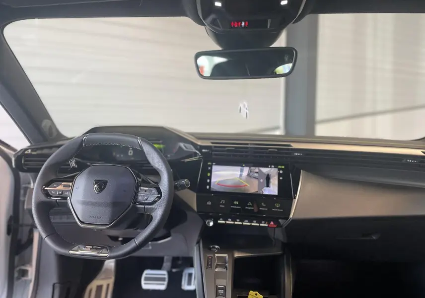 Vue intérieure avant de la Peugeot 308 blanche 2025, volant cuir, écran tactile avec caméra de recul affichée.
