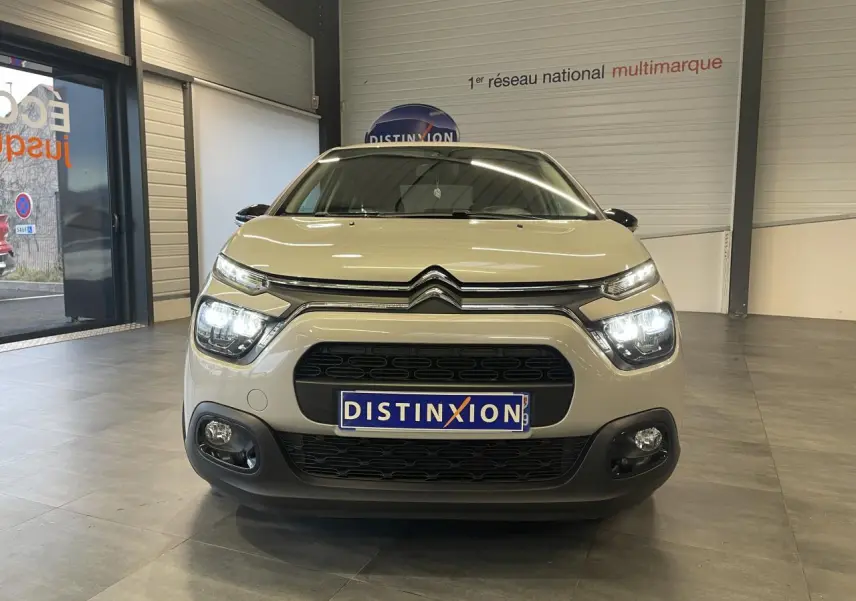 Vue frontale d'une Citroën C3 beige clair 2024 avec feux allumés et calandre noire dans un showroom.