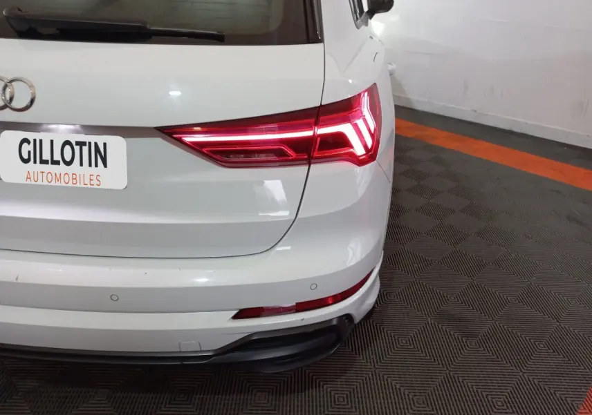 Vue arrière droite d'un Audi Q3 blanc 45 TFSI E S Line avec feux arrière LED allumés dans un garage.
