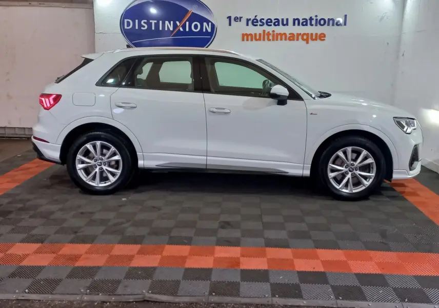 Audi Q3 blanc en vue de profil côté gauche, stationné en intérieur avec jantes alliage et feux arrière allumés.