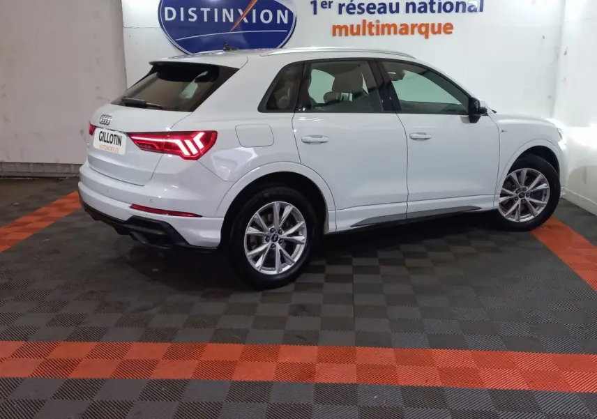 Audi Q3 blanc en vue 3/4 arrière droit, avec feux LED allumés et jantes alliage visibles en intérieur showroom.