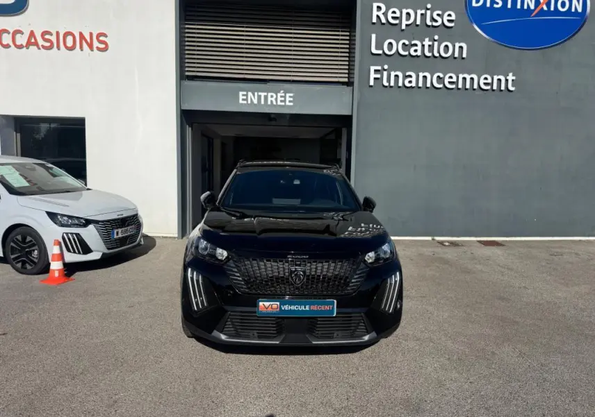 Peugeot 2008 Hybrid noir vue de face, avec barres de toit et calandre distinctive, stationné devant un bâtiment commercial.