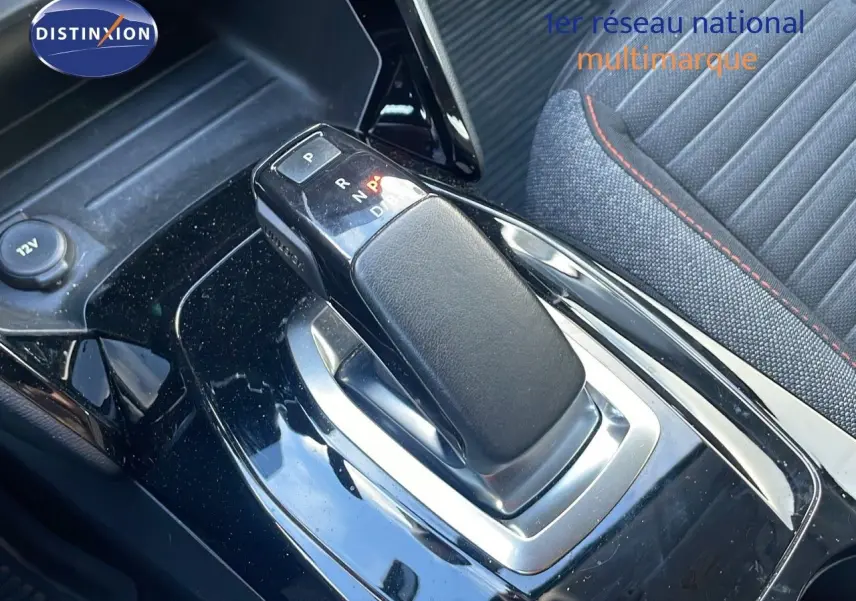 Le levier de vitesse automatique noir brillant de la Peugeot 208 électrique, avec siège tissu gris et surpiqûres rouges visible en arrière-plan.