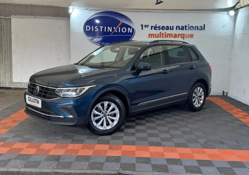 Volkswagen Tiguan bleu en 3/4 avant droit avec rails de toit noirs, stationné sur sol à damiers gris et orange.