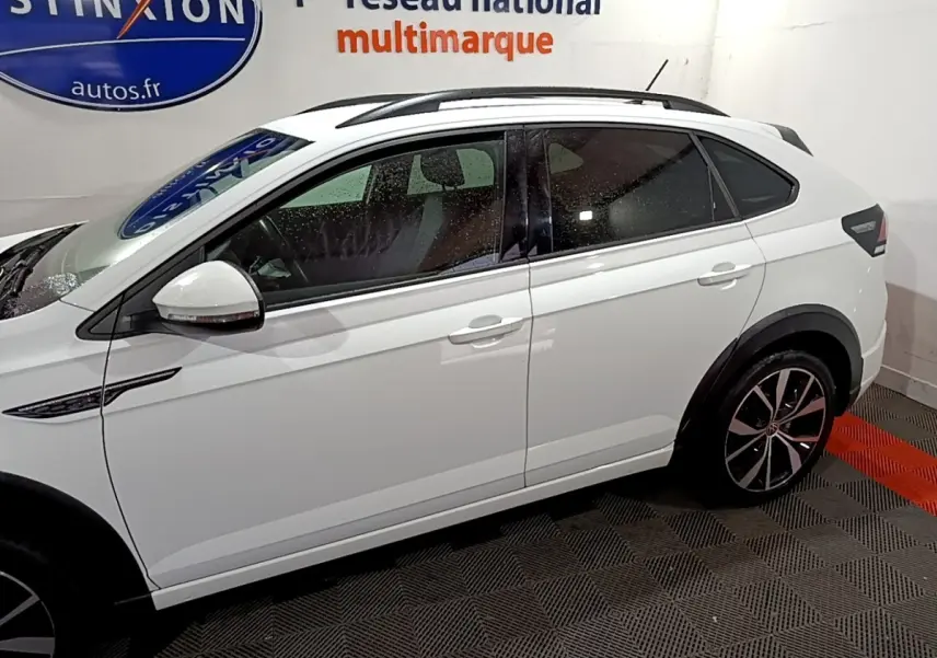 Vue de profil côté gauche d'un Volkswagen Taigo blanc avec jantes alliage et rails de toit noirs en intérieur showroom.