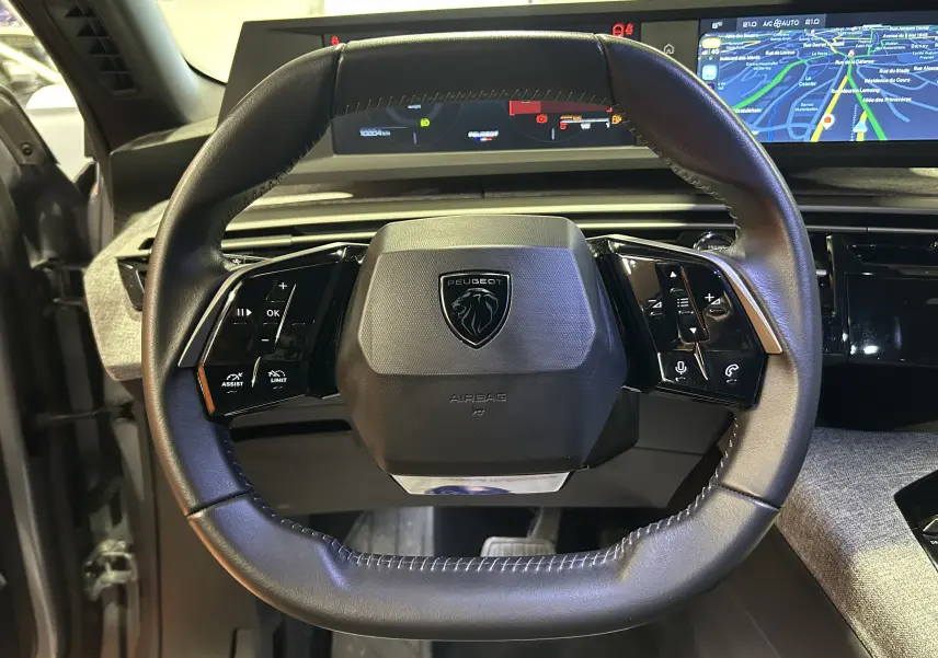 Vue rapprochée du volant cuir noir du Peugeot 5008 III Hybrid 145, avec commandes intégrées et tableau de bord numérique.