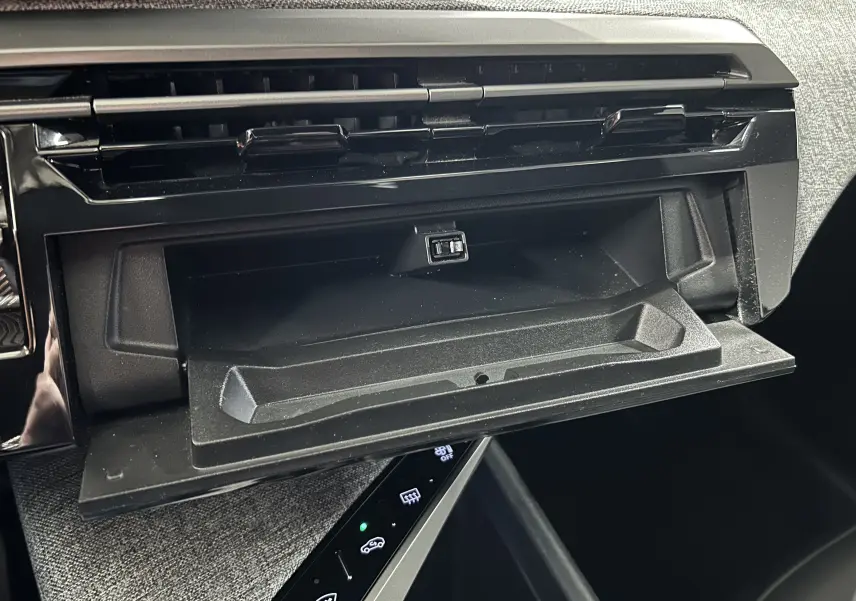 Gros plan sur la boîte à gants ouverte et la console centrale noire du Peugeot 5008 hybride 2025, intérieur gris.