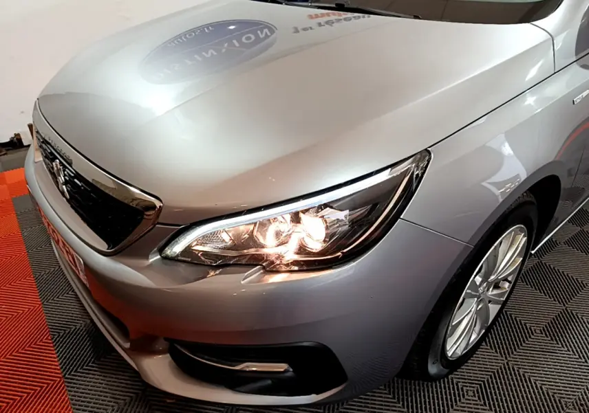 Vue 3/4 avant droit d'une Peugeot 308 gris clair avec phares allumés et jantes alliage visibles.