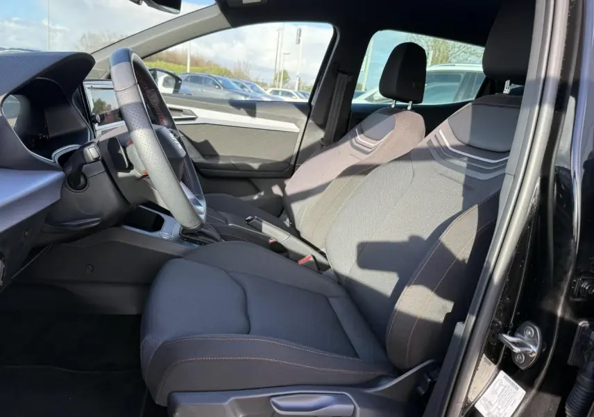 Intérieur de la SEAT IBIZA 2024 vue côté conducteur, sièges tissu gris foncé avec surpiqûres rouges et volant cuir multifonction.