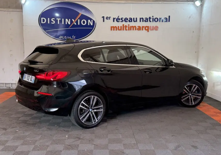 BMW Série 1 116d noir vue 3/4 arrière droit dans un showroom avec logo Distinxion en arrière-plan.
