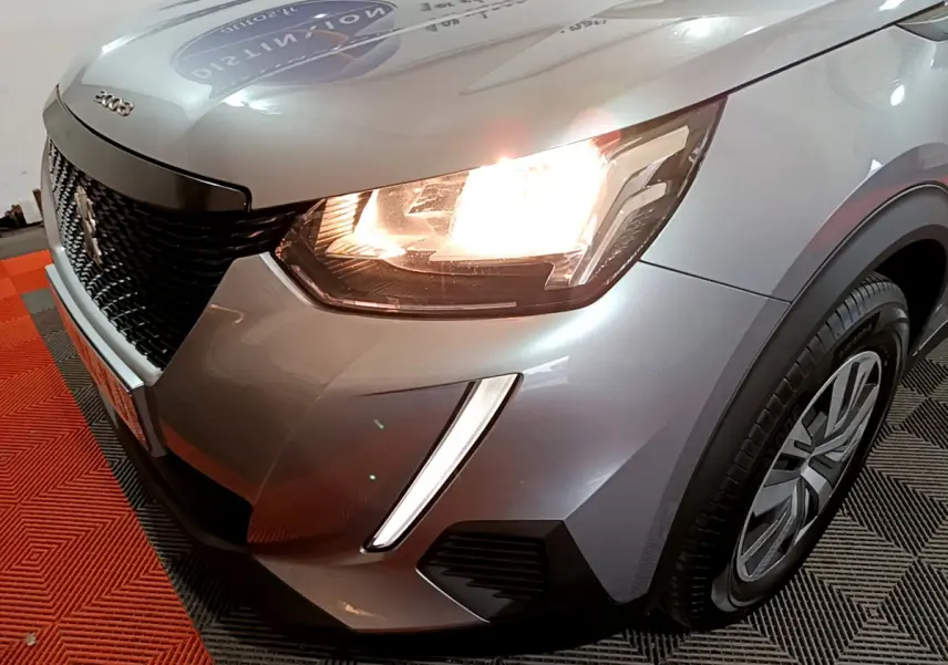 Gros plan sur l'avant droit d'un Peugeot 2008 gris clair 2022 avec phares allumés et calandre noire.