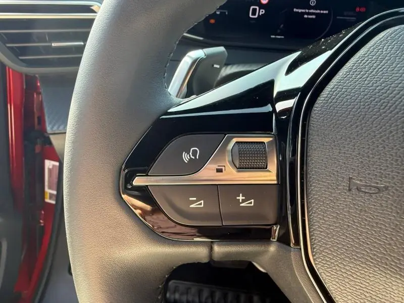 Gros plan sur les commandes gauche du volant du Peugeot 2008 Rouge Elixir, avec tableau de bord numérique en arrière-plan.