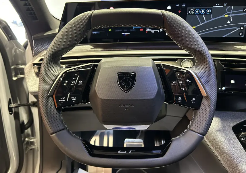 Vue rapprochée du volant GT perforé du Peugeot 3008 Hybrid blanc, avec tableau de bord numérique et commandes intégrées.