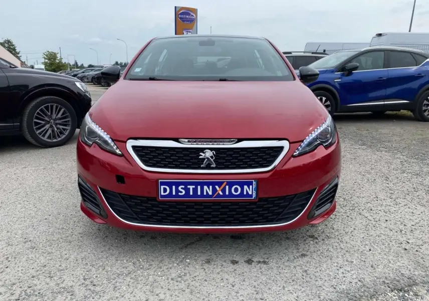 Vue frontale d'une Peugeot 308 GTi rouge avec calandre chromée et phares LED allumés sur parking.