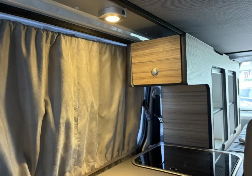 Intérieur du Volkswagen Transporter aménagé, vue latérale droite sur la kitchenette bois clair avec rideau beige fermé.