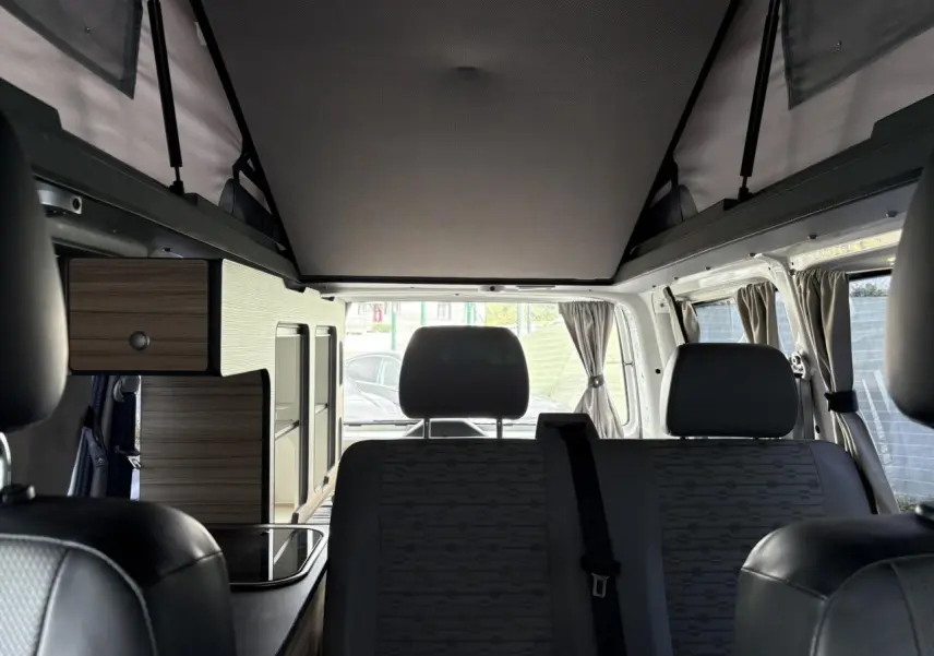 Intérieur du Volkswagen Transporter aménagé, vue arrière montrant sièges gris et rangements bois clairs.