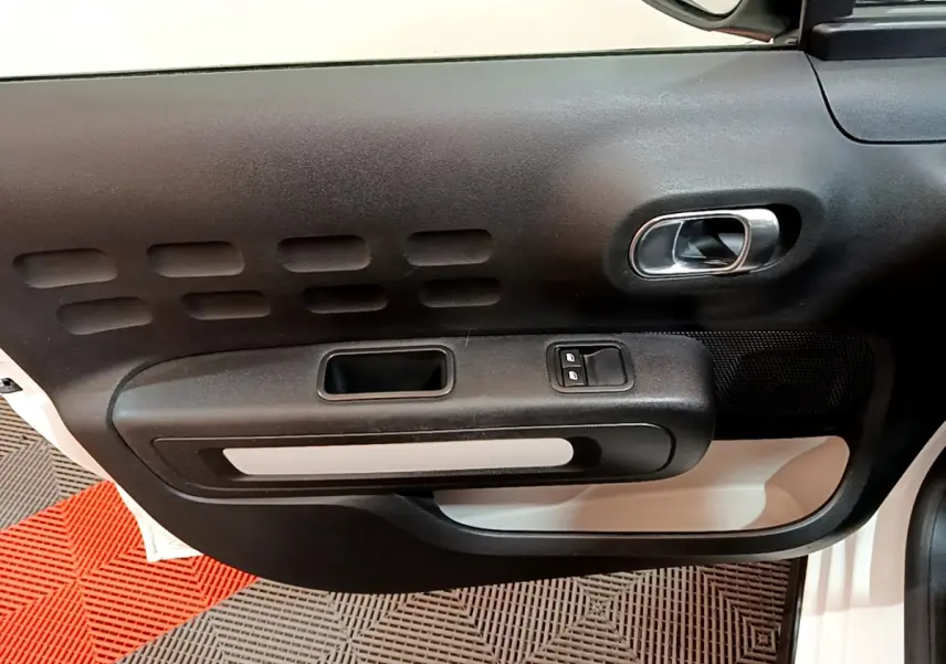 Intérieur de la porte avant gauche d'une Citroën C3 blanche, avec poignée argentée et commandes de vitres électriques.