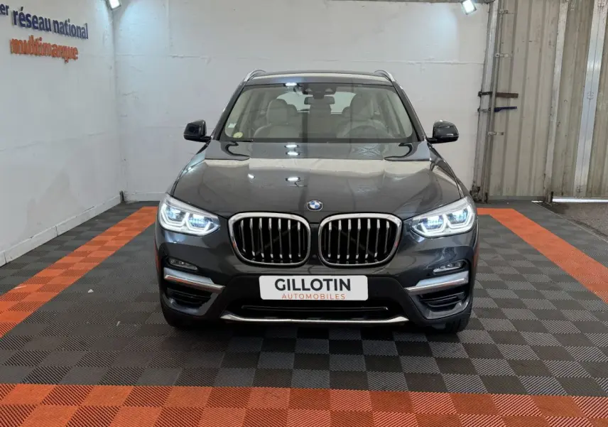 BMW X3 xDrive30d gris foncé vue de face avec phares LED allumés dans un showroom au sol noir et orange.