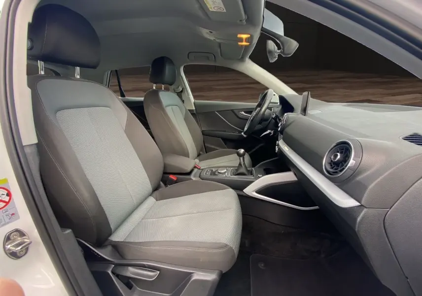 Vue intérieure côté conducteur de l'Audi Q2 blanc 2019, montrant sièges tissu gris et tableau de bord moderne.