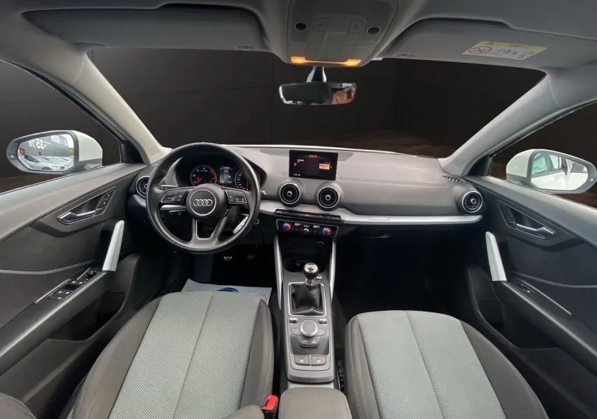 Vue intérieure avant de l'Audi Q2 30 TDI 2019, tableau de bord noir avec volant multifonction et sièges tissu gris.